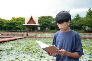 asiatisch Junge lesen Buch beim Garten thailändisch Stil auf Teich und Himmel Sonnenlicht Hintergrund, nah oben Junge Sitzt, Junge Blau Hemd, Kinder mit Buch, Konzept Lernen und Bildung, natürlich schön Hintergrund. foto