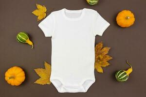 Weiß Baby Mädchen oder Junge Bodysuit eben legen Attrappe, Lehrmodell, Simulation mit Kürbisse und gefallen Blätter auf dunkel Hintergrund. Design Strampler Vorlage, drucken Präsentation spotten hoch. oben Sicht. foto