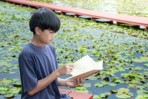 asiatisch Junge lesen Buch beim Flussufer auf Lotus Blume und Sonnenlicht Hintergrund, nah oben Junge Sitzt, Junge Blau Hemd, Kinder mit Buch, Konzept Lernen und Bildung, natürlich schön Hintergrund. foto