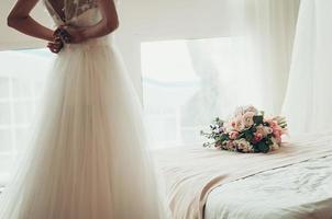 Hochzeitsstrauß auf einem Bett, verschwommene Braut, die ihr Kleid zuknöpft, Rückansicht foto