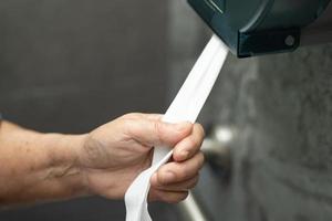 asiatische ältere Patientin zieht das Taschentuch von der Rolle in der Toilette foto