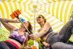 schön Mitte alt Frauen im Sportbekleidung haben Spaß nehmen ein Selfie unter ein Strand Regenschirm foto