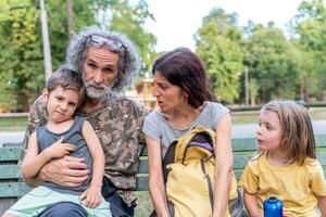 glücklich Familie ist haben Spaß Sitzung auf ein Bank im ein draussen Park foto