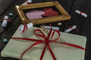 eben legen romantisch Hintergrund. Geschenkbox, gerollt Wunsch Papiere, und hölzern Rahmen mit Papier Herzen auf dunkel Holz Tafel. foto