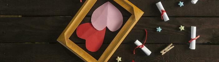 eben legen romantisch Hintergrund. Geschenkbox, gerollt Wunsch Papiere, und hölzern Rahmen mit Papier Herzen auf dunkel Holz Tafel. foto