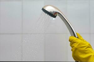 ein Person im Gummi Handschuhe halten Duschkopf mit fließend Wasser. Frauen im Gelb schützend Handschuhe halten das Kopf von das Dusche mit Laufen Wasser im das Bad foto