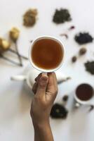 Hand halten Tee Tasse. Frau halten ein Tasse von Tee mit Tee Topf, getrocknet Rose Blumen und andere Tee Zutaten auf das Hintergrund. foto