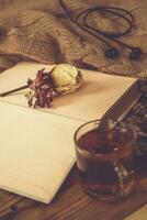fallen Hintergrund. Herbst Komposition mit heiß Tee, Buch, trocken Rose Blume und gestrickt Pullover. ein Tasse von heiß Tee, gestrickt Schal auf hölzern Tisch. foto