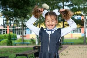 heiter komisch Mädchen mit ein zahnlos Lächeln im ein Schule Uniform mit Weiß Bögen im Schule Hof. zurück zu Schule, September 1. ein glücklich Schüler. primär Ausbildung, elementar Klasse. Porträt von ein Schüler foto