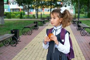 Mädchen mit ein Rucksack Essen Kuchen in der Nähe von Schule. ein schnell Snack mit ein Brötchen, ungesund Essen, Mittagessen von Schule. zurück zu Schule. Ausbildung, primär Schule Klassen, September 1 foto