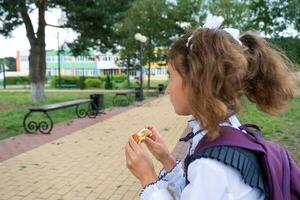 Mädchen mit ein Rucksack Essen Kuchen in der Nähe von Schule. ein schnell Snack mit ein Brötchen, ungesund Essen, Mittagessen von Schule. zurück zu Schule. Ausbildung, primär Schule Klassen, September 1 foto