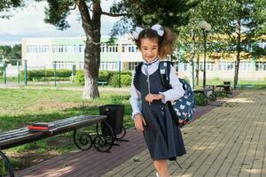 heiter komisch Mädchen mit ein zahnlos Lächeln im ein Schule Uniform mit Weiß Bögen im Schule Hof. zurück zu Schule, September 1. glücklich Schüler mit ein Rucksack. primär Ausbildung, elementar Klasse. foto