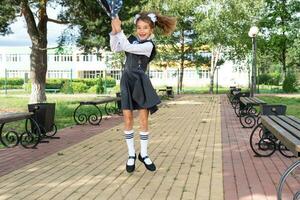 heiter komisch Mädchen mit zahnlos Lächeln im Schule Uniform mit Weiß Bögen im Schule Hof wirft das Rucksack. zurück zu Schule, September 1. glücklich Schüler. primär Ausbildung, elementar Klasse. foto