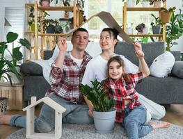 traditionell Familie mit Kind sind genießen Neu heim, Sitzung auf das Sofa und halten das Dach. Hypothek, Versicherung und Schutz, Kauf und ziehen um in Ihre besitzen heim, Grün Haus mit eingetopft Pflanze foto