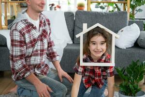 unvollständig Familie Papa und Tochter sind genießen Neu heim, Sitzung auf Sofa. Hypothek, Versicherung und Schutz, Kauf und ziehen um in Ihre besitzen heim, Grün Haus mit eingetopft Pflanze foto