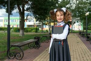 Mädchen mit Rucksack, Schule Uniform mit Weiß Bögen und Stapel von Bücher in der Nähe von Schule. zurück zu Schule, glücklich Schüler, schwer Lehrbücher. Ausbildung, primär Schule Klassen, September 1 foto