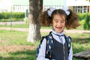 heiter komisch Mädchen mit ein zahnlos Lächeln im ein Schule Uniform mit Weiß Bögen im Schule Hof. zurück zu Schule, September 1. glücklich Schüler mit ein Rucksack. primär Ausbildung, elementar Klasse. foto