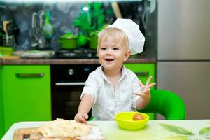 glücklich wenig Junge vorbereiten Teig im Küche beim Tisch. Dort sind Teig Produkte auf Tisch, gekleidet wie Koch foto