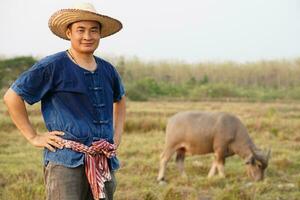 gut aussehend asiatisch Mann Farmer trägt Hut, Blau Shirt, stellen Hände auf Taille, steht beim Tier Bauernhof. Konzept, Vieh.Thai Bauern erziehen und nehmen Pflege Büffel wie wirtschaftlich und Export Tiere. foto