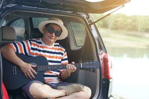 asiatisch Mann Reisender sitzen und abspielen das Gitarre auf Kofferraum von Auto Konzept, entspannen Zeit. Urlaub, Urlaub oder Wochenende Ausflug durch Privat Fahrzeug. Transport. Straße reise.geben Zeit zum du selbst. genießen singen Lieder foto