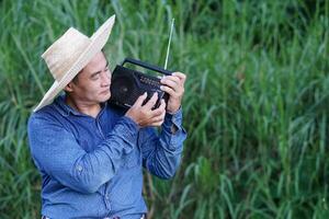 asiatisch Mann Farmer trägt Hut, Blau Shirt, hält Transistor Radio auf seine Schulter beim Garten, fühlt sich glücklich. Konzept, glücklich Arbeiten entlang mit Musik, Nachricht, Information von Radio. Land Lebensstil. foto