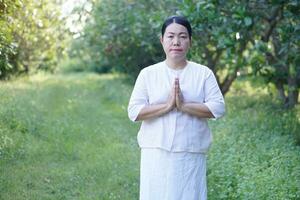 asiatisch Frau Buddhist trägt Weiß Kostüm, stellen Palmen zusammen von beten, steht beim still Wald. Konzept, Kultur von Glaube. religiös Aktivität. Frieden von Verstand verwalten foto