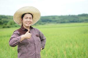 asiatisch Frau Farmer trägt Hut, Daumen hoch, fühlt sich zuversichtlich beim Paddy Feld. Konzept, Landwirtschaft Beruf. thailändisch Bauer. Arbeiten mit Natur. organisch Landwirtschaft. foto