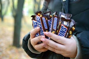 Charkow, Ukraine - - Oktober 8, 2019 ein jung kaukasisch Frau zeigt an viele kichert Schokolade Riegel im braun Verpackung im Herbst Park. kichert Schokolade hergestellt durch Mars foto