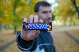 Charkow, Ukraine - - Oktober 8, 2019 ein jung kaukasisch bärtig Mann zeigt an kichert Schokolade Bar im braun Verpackung im Herbst Park. kichert Schokolade hergestellt durch Mars foto