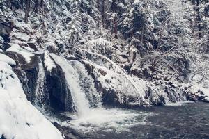 Winterwasserfall zwischen Wäldern und Bergen mit Schnee bedeckt foto