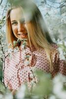 blond Mädchen auf ein Frühling gehen im das Garten mit Kirsche blüht. weiblich Porträt, Nahansicht. ein Mädchen im ein Rosa Polka Punkt Kleid. foto