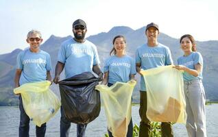 multiethnisch Menschen im Blau Freiwillige T-Shirt Freiwillige halten Abfall Sortierung Plastik Tasche Stehen durch das Fluss, ein vielfältig Gruppe von Freiwillige beitreten zusammen zu Aufräumen das Sand Strand zusammen foto