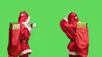 pünktlich Santa prüft Zeit auf Armbanduhr während er trägt rot Sack gefüllt mit die Geschenke, tragen festlich Charakter Kostüm auf grüner Bildschirm. Heilige Nick warten mit groß Tasche von Geschenke und Spielzeuge. foto