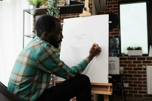 jung afrikanisch amerikanisch Kerl ausdrücken selbst durch Kunst, Teilnahme Zeichnung Klassen, männlich Schüler Verbesserung Skizzierung Fähigkeiten. talentiert Künstler Sitzung beim Staffelei Erstellen Meisterstück auf Segeltuch foto