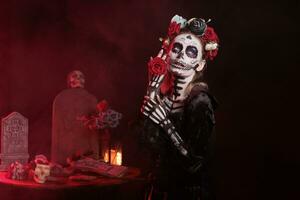 Dame von Tod halten Rosen auf Mexikaner Urlaub, suchen mögen Santa muerte mit Halloween Kostüm und Schädel machen hoch. Schauspielkunst gruselig und Glanz auf Tag von das tot Feier im Studio. foto