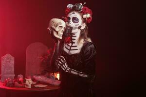 la Kavalera catrina Schauspielkunst gruselig mit Schädel im Hand, halten schwarz Rosen und tragen unheimlich machen hoch. suchen mögen Santa muerte mit Blumen Krone und Körper Kunst auf heilig Mexikaner Feier. foto