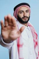 zuversichtlich Muslim Mann Herstellung verboten halt Zeichen mit Palme und suchen beim Kamera. ernst arabisch Person gekleidet im traditionell Kleider zeigen Ablehnung Geste mit Hand Studio Porträt foto