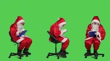 Winter Charakter nehmen Anmerkungen auf Papiere, Sitzung auf Stuhl zu schreiben Information auf Zwischenablage während Weihnachten Vorabend Urlaub. Santa claus Mann im Cosplay posieren Über grüner Bildschirm Hintergrund. foto