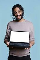 heiter arabisch Mann Stehen mit Laptop zeigen leer Bildschirm zum Netz Seite spotten oben Porträt. Person halten tragbar Computer mit Weiß Anzeige zum Werbung und suchen beim Kamera foto