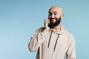 heiter arabisch Mann haben Spaß Konversation auf Smartphone und lächelnd während reden. Person chatten auf Handy, Mobiltelefon Telefon und Hören zu Gesprächspartner mit froh aufgeregt Gesichts- Ausdruck foto