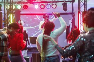 dj Sprechen zu Publikum im Mikrofon während durchführen auf Bühne im Nachtclub. Menschen Tanzen und erziehen Hände auf Tanzfläche während Teilnahme elektronisch Musik- Konzert im Verein foto
