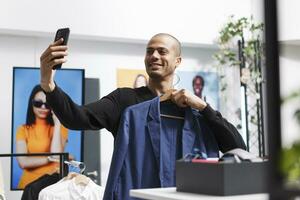 lächelnd arabisch Mann halten Aufhänger mit stilvoll Jacke während nehmen Selfie mit Smartphone Vorderseite Kamera im Kleidung speichern. Blogger präsentieren bekleidung zu Sozial Medien Kanal Anhänger im Boutique foto