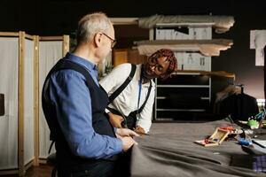 hilfreich alt Meister Schneider führen glücklich Lehrling mit Schneiden Textil- Material zum bevorstehende maßgeschneidert sartorial Sammlung. Couturiers im Schneiderei Studio Arbeiten auf Herstellung stilvoll Mode Kleidung foto