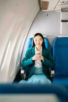 attraktiv asiatisch weiblich Passagier von Flugzeug Sitzung im komfortabel Sitz während Arbeiten Laptop und Tablette mit spotten oben Bereich mit kabellos Verbindung. Reise im Stil, Arbeit mit Anmut. foto