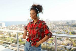 schließen oben Porträt von amerikanisch Frau genießen schön Musik- durch Kopfhörer , gekleidet im kariert Shirt, Stehen auf Dach. städtisch Landschaft Hintergrund. foto