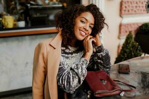 draussen Porträt von schön lächelnd schwarz Frau mit stilvoll afro Haare Sitzung im Cafe im Paris. tragen modisch Samt Kleid und Beige Mantel. Luxus Tasche. foto