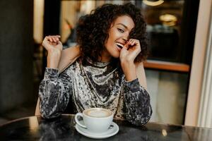 schließen oben Lebensstil Porträt von glücklich sorglos schwarz Frau genießen Kaffee brechen im Paris. tragen elegant Samt Kleid. perfekt Weiß Zähne. lockig Haare. foto