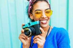 Lachen schwarz Mädchen mit süß Frisur halten retro Kamera und suchen beim Kamera. draussen schließen oben Schuss von glücklich Fotograf. Gelb Sonnenbrille. foto