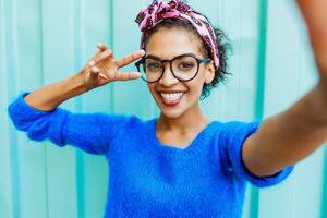 schön afrikanisch Mädchen mit stilvoll Frisur Herstellung selbst Porträt durch Kamera auf Türkis Hintergrund. zeigen Zeichen durch Hand. positiv Stimmung. Hipster Brille. foto