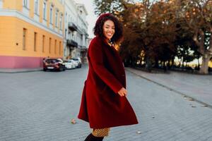 lächelnd schwarz Mädchen im tolle Winter Outfit und Zubehör posieren auf Straße Hintergrund. wellig Haare. flauschige Pullover. foto